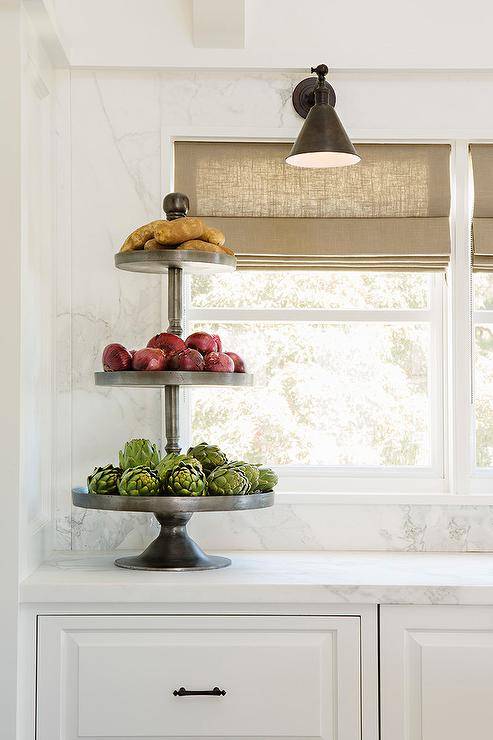 Beige roman shades hang from window framed by a honed white marble backsplash lit by an oil rubbed bronze swing arm sconce. Beneath the windows, white raised panel cabinets are adored with oil rubbed bronze pulls and a honed white marble countertop topped with a vintage tiered fruit tray.