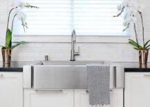 White shaker cabinets fitted with nickel pulls are contrasted with a black countertop and hold a stainless steel apron sink with a satin nickel gooseneck faucet beneath a window dressed in white blinds layered under a black and white roman shade.