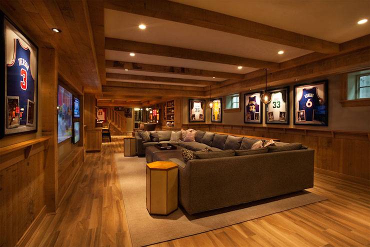 Basement man cave features wood ceiling beams interspersed with pot lighting over top half of walls painted beige and bottom half of walls clad in rustic paneling highlighting framed basketball jerseys illuminated by iron and glass globe light pendants. A U shaped gray sectional sofa is paired with side-by-side brown leather ottoman coffee tables as well as hex accent tables atop sand colored rug facing a flatscreen TV.