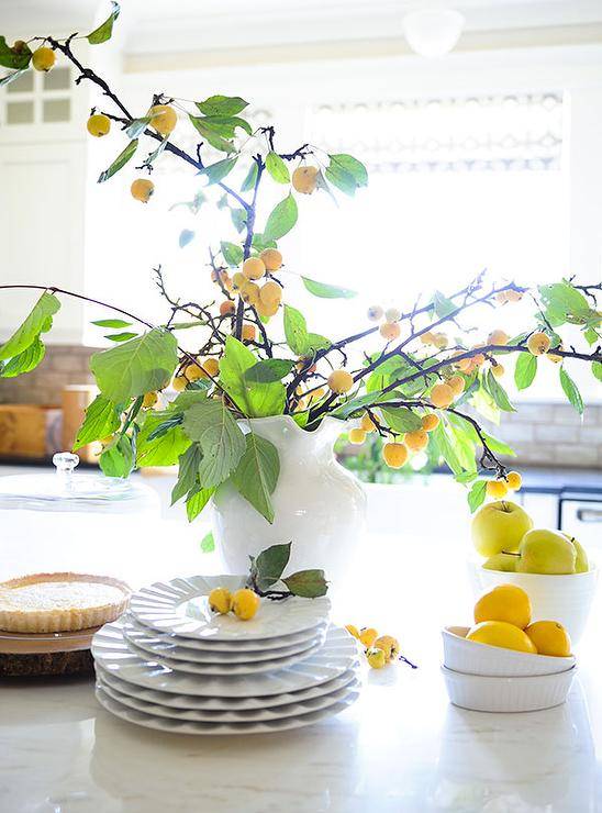 White jug with fruit branches accents a marble countertop in a transitional kitchen with lovely natural light.
