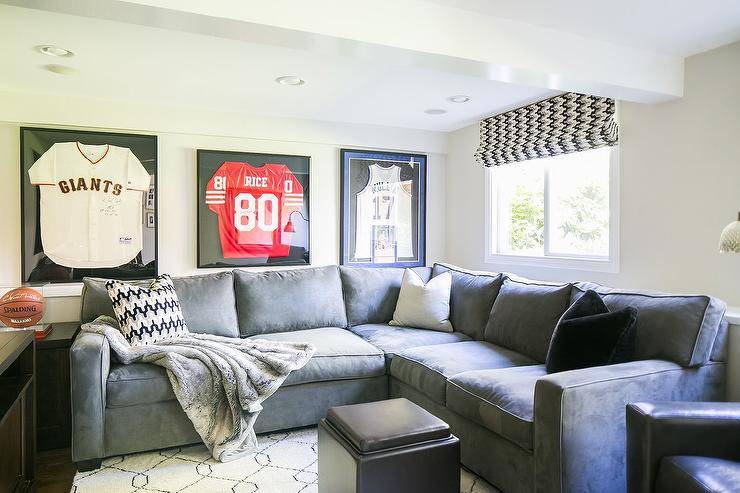Framed sports jerseys hang above a gray microfiber sectional placed on a white and black Moroccan wool rug beneath a window covered in a white and black chevron roman shade.