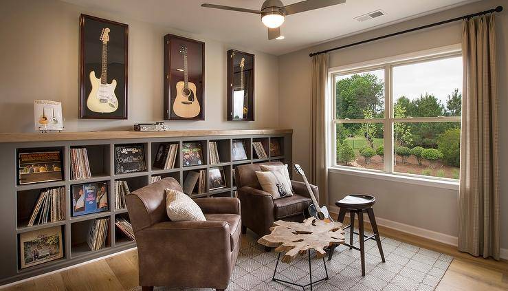 Man cave and music room combo features a row of guitars framed in shadow boxes placed over an industrial shelving unit filled with vintage vinyl records. Industrial man cave room boasts a pair of brown leather club chairs facing a driftwood coffee table placed in front of windows dressed in beige curtains.