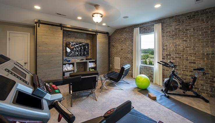 Industrial man cave and exercise room combo features workout equipment and yoga mats placed in front of exposed brick walls. Man cave boasts a pair of black tufted accent chairs, Barcelona Chairs, facing a flat panel tv and workout accessories concealed behind double sliding doors on rails.