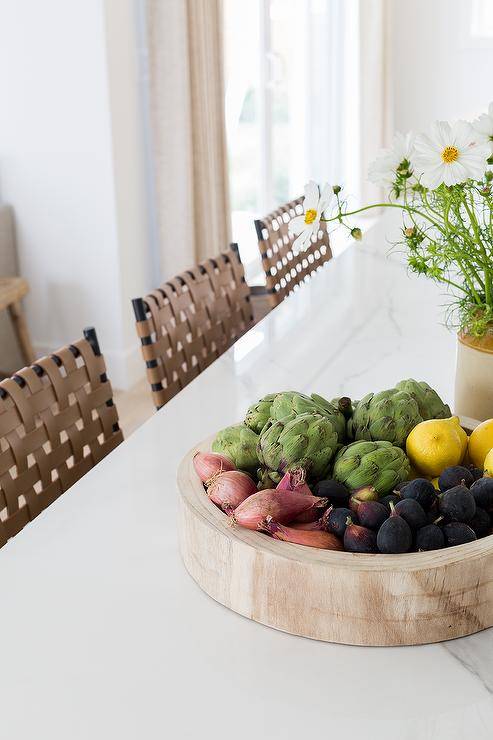 A mango wood fruit bowl sits on a marble island countertop fixed in front of brown woven leather stools.