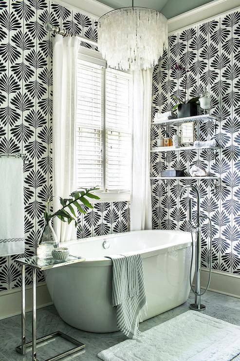 An oval soaking bathtub sits on gray floor tiles behind a white bath mat and is paired with a nickel and glass c-table and a polished nickel floor mount tub filler. The tub is positioned beneath a window fitted with white blinds layered behind white curtains accenting a wall covered in Clarke & Clarke Leaf Foliage Vine Wallpaper. A capiz chandelier hangs over the tub and in front of stacked marble shelves.
