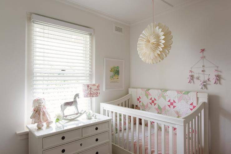 Beneath a window, a white dresser sits beneath a window covered in blinds in a gorgeous white nursery. A white crib dressed in pink and green bedding is paired with a teddy bear mobile and placed under a framed art piece.