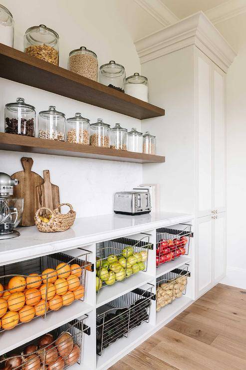 Kitchen pantry features stacked pull out fruit and vegetable bins under floating shelves with vintage jars and white on white pantry cabinets.