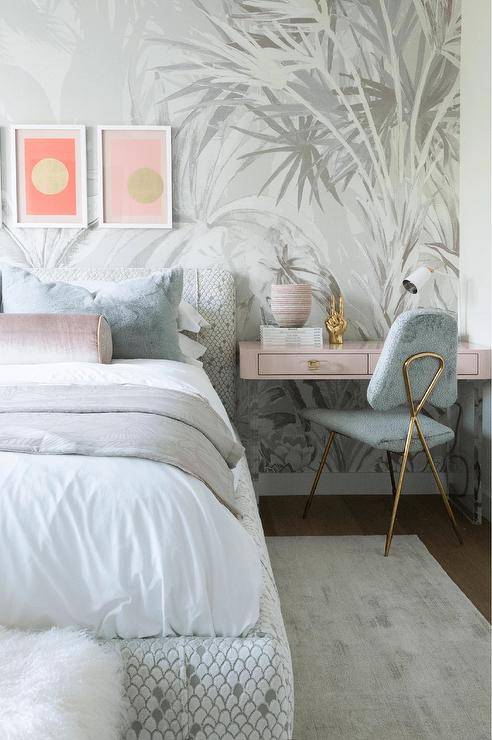 Bedroom features a pink wall mount desk as a bedside table with a gray chair and a white and gray bed with a pink velvet bolster pillow.