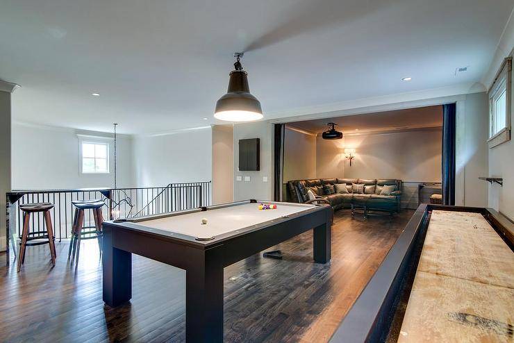 Ideal for entertainers, this game room is designed on a second floor in a large home in a darker rugged wood theme. Black industrial flush mount lights hang over a large landing pool table and movie room. Long shuffleboard is a highlight in this game room adding to the textural wood accents such as a dark stained plank wood floor. A gray curved leather sectional offers comfortable seating in a tv room while the game room displays industrial styled barstools.