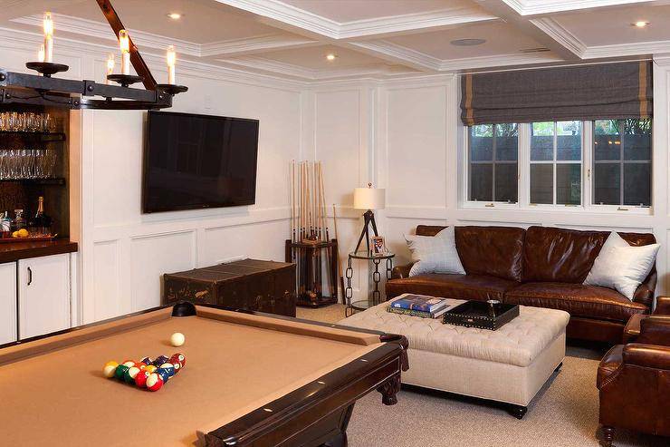Fantastic basement features a coffered ceiling accented with a leather and iron chandelier illuminating a pool table facing a built-in bar nook. Basement man cave boasts a brown leather sofa and chairs facing a beige tufted storage ottoman facing a vintage trunk under a flatscreen TV.