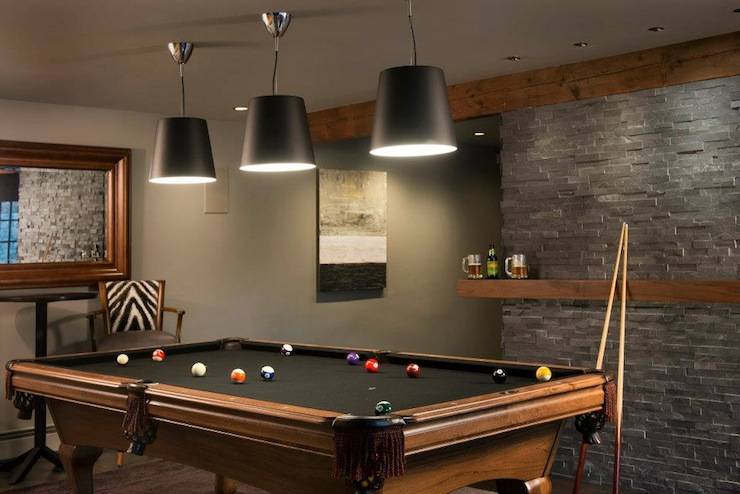 Enviable games room with pool table lit by three black cone shaped pendants. The back wall is covered in a charcoal gray stacked stone accented with a walnut drinks ledge. The remaining walls and ceiling are painted a dark taupe. A small bar height table and bar chair with zebra print back sit at the far end of the pool table with a large framed mirror hanging overhead.