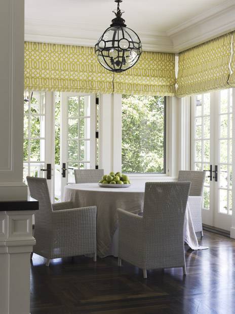Sunshine filled dining room with dark wood floors, picture windows and French doors. The doors and windows are dressed in roman blinds with Kelly Wearstler Imperial Trellis Citrine fabric. The round skirted dining table pairs with four contemporary woven dining chairs. A sphere shaped glass and iron pendant hangs over the round table.
