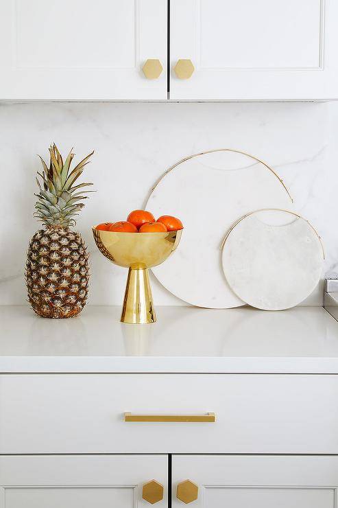 Round marble cutting boards with brass trim decorate a white kitchen atop a white quartz countertop with matching backsplash. Gold hex knobs and a horizontal pull decorate the white shaker cabinets in the most stylish way keeping up with the trends. A gold pedestal fruit bowl highlights out the gold hardware and accents trimming.