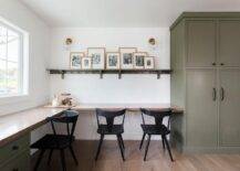 Beautifully appointed homework room features an l-shaped green desk with a wood top seating black vintage desk chairs. A green wooden picture shelf is lit by glass and brass sconces and mounted beside floor-to-ceiling green cabinets.