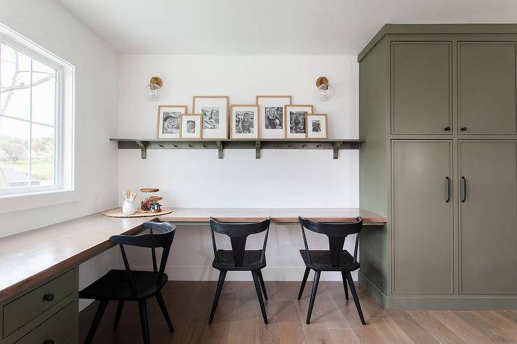 Beautifully appointed homework room features an l-shaped green desk with a wood top seating black vintage desk chairs. A green wooden picture shelf is lit by glass and brass sconces and mounted beside floor-to-ceiling green cabinets.