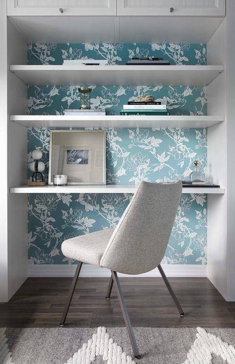 A heather gray desk chair sits on a gray rug in front of a white floating desk fitted in a nook beneath styled white floating shelves mounted under white cabinets to a wall covered in Graham & Brown Jiao Blue Wallpaper.