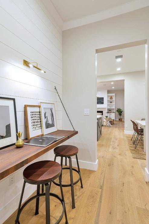 Cottage style workspace designed with a shiplap wall displaying a wooden desk with a metal chain decorated with framed wall art. Two round wood and metal stools add seating to the workspace perfect for the cottage appeal.