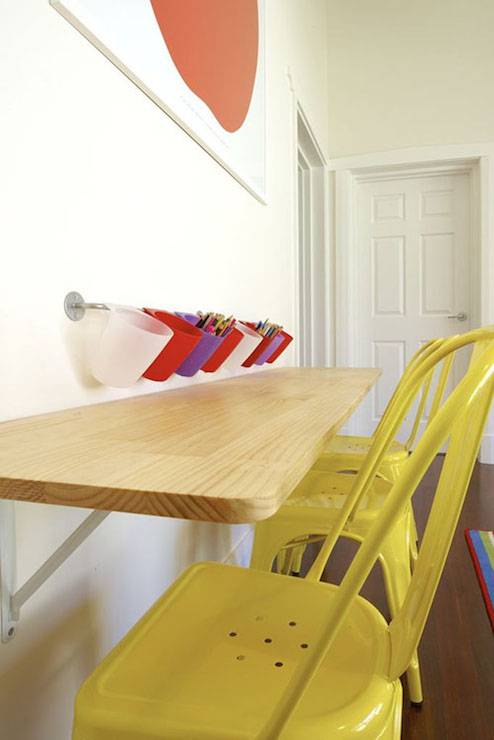 Kids' playroom with long wall mounted wooden desk lined with kid sized yellow Tabouret chairs with wall mounted pencil holders above.