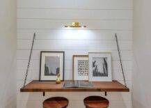 Workspace in a cottage nook features a brass picture light above a wood desk with metal brackets and chains completed with backless stools for practical storing options.