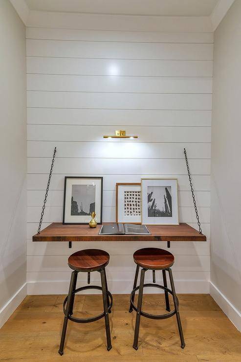 Workspace in a cottage nook features a brass picture light above a wood desk with metal brackets and chains completed with backless stools for practical storing options.