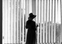 Black and white photo of a woman standing in front of a window with vertical blinds.