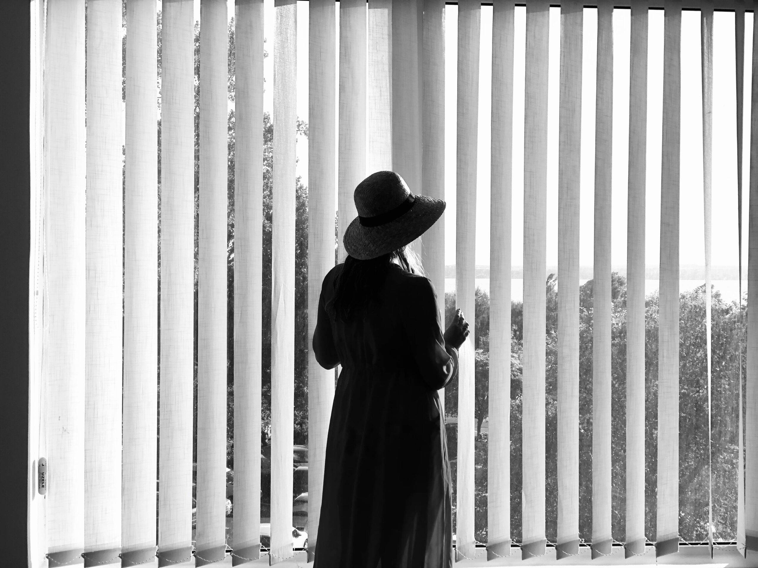 black and white photo of a woman standing in front of a window with vertical blinds