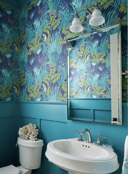 Blue powder room with blue wainscoting features an oval pedestal sink under a beveled mirror mounted on blue and green floral wallpaper.