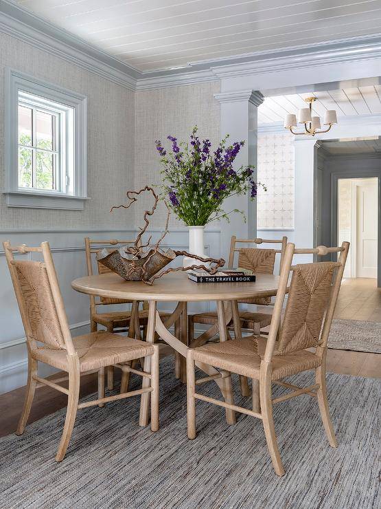 Placed around a round beige wooden dining table, beige woven dining chairs sit on a gray rug in front of gray wallpapered walls finished with light blue wainscoting and light blue crown moldings and baseboards. The ceiling is completed with glossy white shiplap.