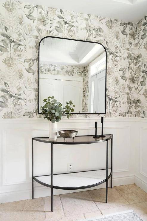 An oil rubbed bronze arch mirror is mounted on gray floral wallpaper over a half moon bonze console table on wainscoting.
