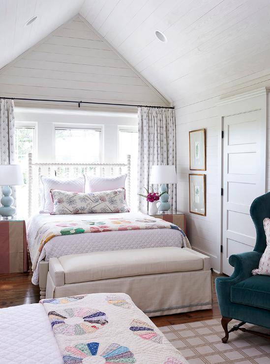 Farmhouse style bedroom displaying a bed in front of a window under a vaulted shiplap ceiling with white skirted bedside tables displaying baby blue triple gourd lamps. Beautiful country furnishings and decor charm this bedroom staring with a tan linen bench at the foot of the bed and curtains behind the bed.