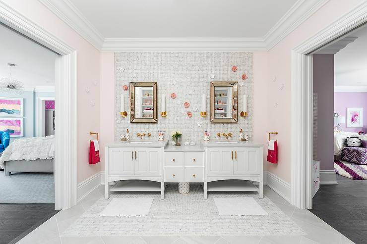 White and pink Jack and Jill girl's bathroom is illuminated by Kohler Purist Single Sconces mounted on white mosaic marble grid tiles on either side of beveled beaded mirrors fixed above a white dual washstand. The washstand is fitted with white cabinets adoring brass hardware, a white quartz countertop, and Kohler Purist Wall Mount Faucets fixed above oval sinks positioned flanking drop-down middle drawers. Side walls are painted in pale pink with the flower is covered in white mosaic marble tiles framed by large white marble tiles.