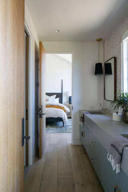 Jack and Jill galley style bathroom design features a blue double washstand with gray quartz countertop and light wooden floors that lead to a bedroom that boasts a black poster bed with white and orange bedding.