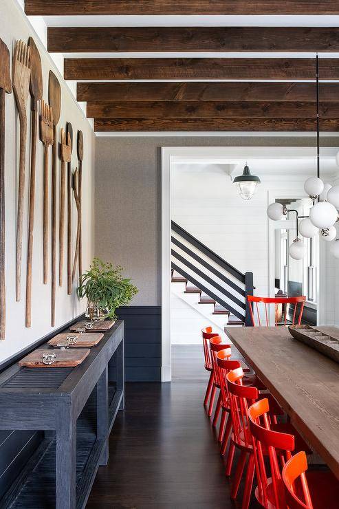 Large forks and spoons decorate a rustic farmhouse dining room wall along with a black long buffet table. Red spindle dining chairs surround a light wood dining table illuminated by a sputnik globe chandelier hanging from wooden ceiling beams.