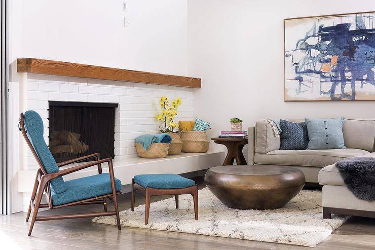 Wonderfully designed living room features a large blue abstract art piece hung above a gray shelter back sectional topped with blue pillows and placed facing a round brass coffee table positioned on a Moroccan trellis rug. A brown and blue mid-century modern chair is placed at an angle in front of a white brick mid-century modern fireplace finished with a stained wood mantel.
