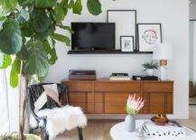 Contemporary living room features a black picture ledge over a mid century modern TV console, a black accent chair with white faux fur and a round gold coffee table.