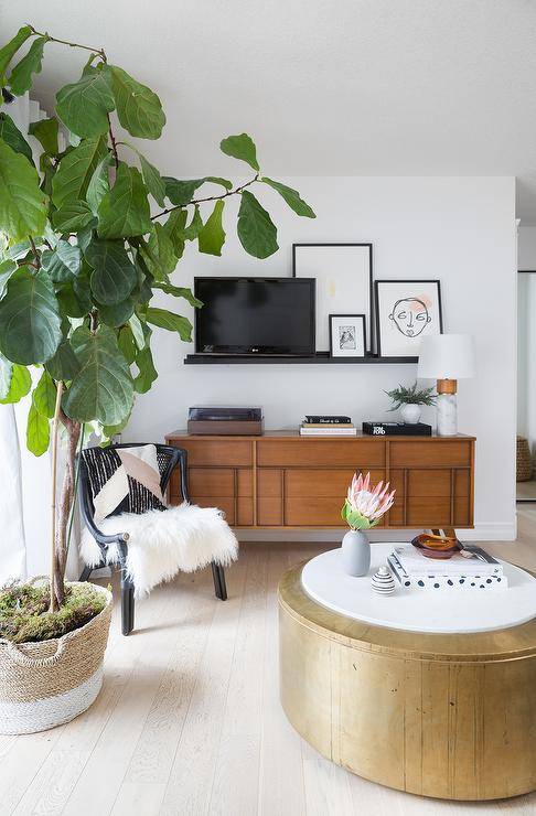 Contemporary living room features a black picture ledge over a mid century modern TV console, a black accent chair with white faux fur and a round gold coffee table.