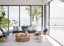 Brown and blue mid century modern lounge chairs on gray tiles join a round wood and rope metal coffee table in a sunroom with glass doors and windows.