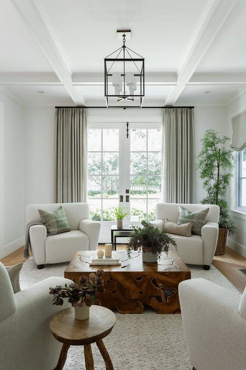 Living room features sage green pillows on light gray accent chairs, a burl wood coffee table atop a light gray rug, a round wooden stool as accent table and a tall potted plant.