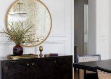 A round gold mirror is mounted on wainscoting over a dark brown buffet cabinet.