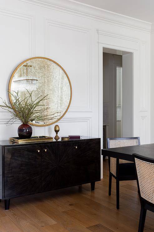 A round gold mirror is mounted on wainscoting over a dark brown buffet cabinet.