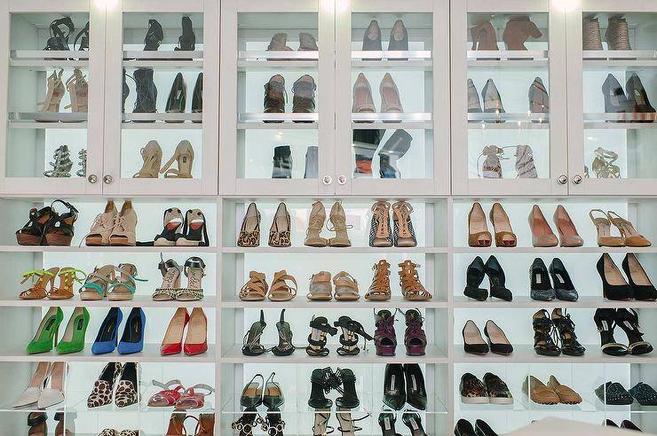 This walk in closet wall is lined with white shoe shelves positioned between glass front shoe cabinets and acrylic shoe cubbies.