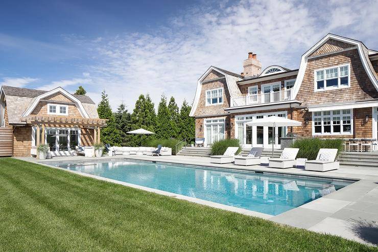 Cape cod cottage features light brown shake siding, white pool loungers at an in ground pool and an outdoor umbrella.