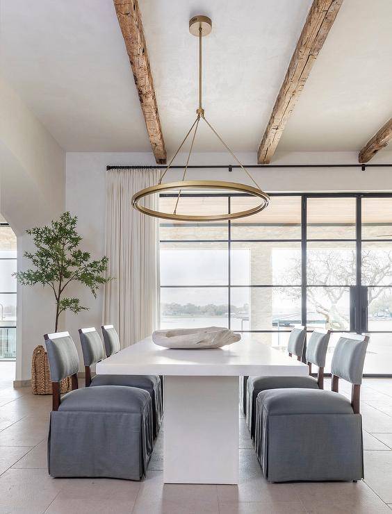 A brass ring chandelier with wooden ceiling beams illuminates a white pedestal dining table with gray skirted slipper chairs.