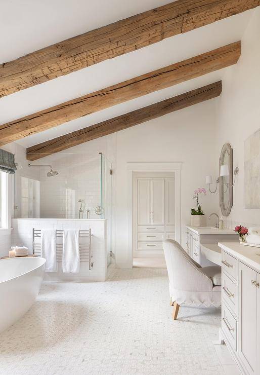 Rustic wood beams accent a sloped bathroom ceiling and are fixed above an oval freestanding bathtub positioned under a window to the side of a towel warmer mounted against a shower.