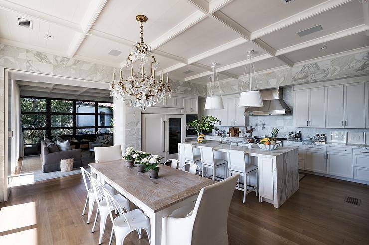 Large, gorgeous open concept kitchen into a dining room featuring a white two-tone farmhouse dining table with white Tolix chairs and natural linen end chairs. A French crystal chandelier drapes over the modern farmhouse table from stunning coffered ceilings.