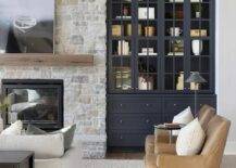 Black built-in display cabinets are fixed in a country style living room beside a gray stone fireplace finished witha hearth and a chunky brown wooden mantel mounted beneath an art piece.