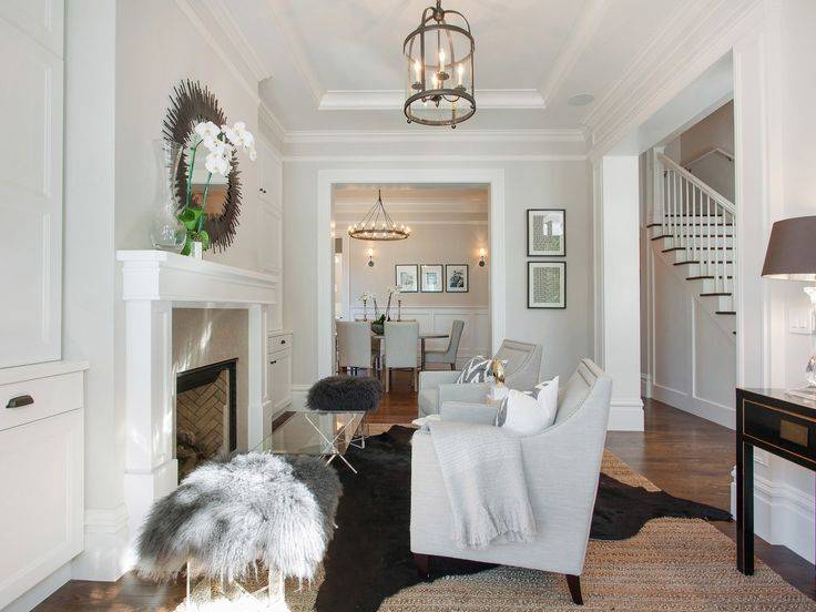 Chic formal living room features a Round Edwardian Entry Lantern illuminating a pair of gray chairs with brass nailhead trim adorned with gray chevron pillows atop a black cowhide rug layered atop a jute rug facing a traditional fireplace. A pair of contemporary lucite stools topped with gray sheepskin flank an x-based coffee table with glass-top stands in front of a fireplace with taupe stone surround situated under an oval metal mirror.