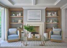 Morgan Harrison Home - A framed art piece hangs from a gray wall over a limestone fireplace flanked by built-in styled oak shelves. A glass and rope coffee table is placed on a white and blue striped rug in front of accent chairs with blue cushions.