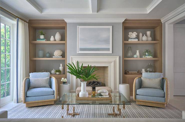 Morgan Harrison Home - A framed art piece hangs from a gray wall over a limestone fireplace flanked by built-in styled oak shelves. A glass and rope coffee table is placed on a white and blue striped rug in front of accent chairs with blue cushions.