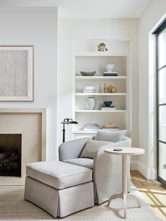 A sconce lights an alcove bookcase built-in behind a dove gray living room chair complemented with a matching ottoman and a round wooden accent table illuminated by a bronze pharmacy floor lamp.